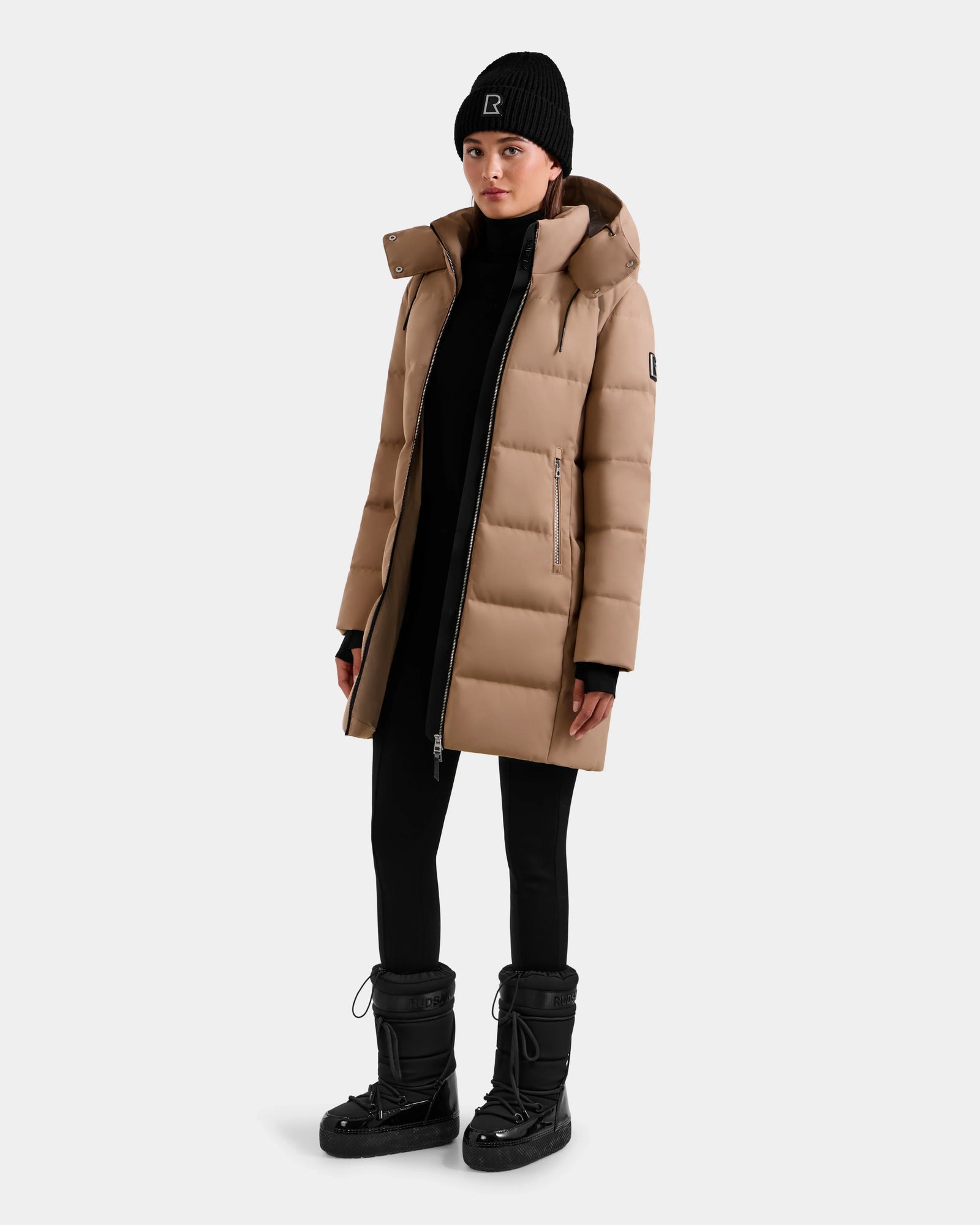A woman wears the Rudsak MIRIAM - L123544 Sand down puffer coat, paired with a black beanie, black leggings, and black winter boots as she stands against a plain light background.