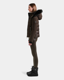 A woman stands in profile wearing the Rudsak MIRRY M - L124533 Espresso women's quilted down coat with a fur-trimmed hood, olive leggings, and black winter boots against a plain light background.