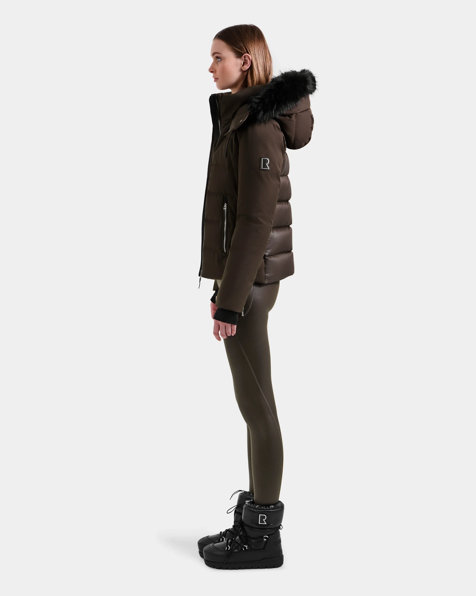 A woman stands in profile wearing the Rudsak MIRRY M - L124533 Espresso women's quilted down coat with a fur-trimmed hood, olive leggings, and black winter boots against a plain light background.