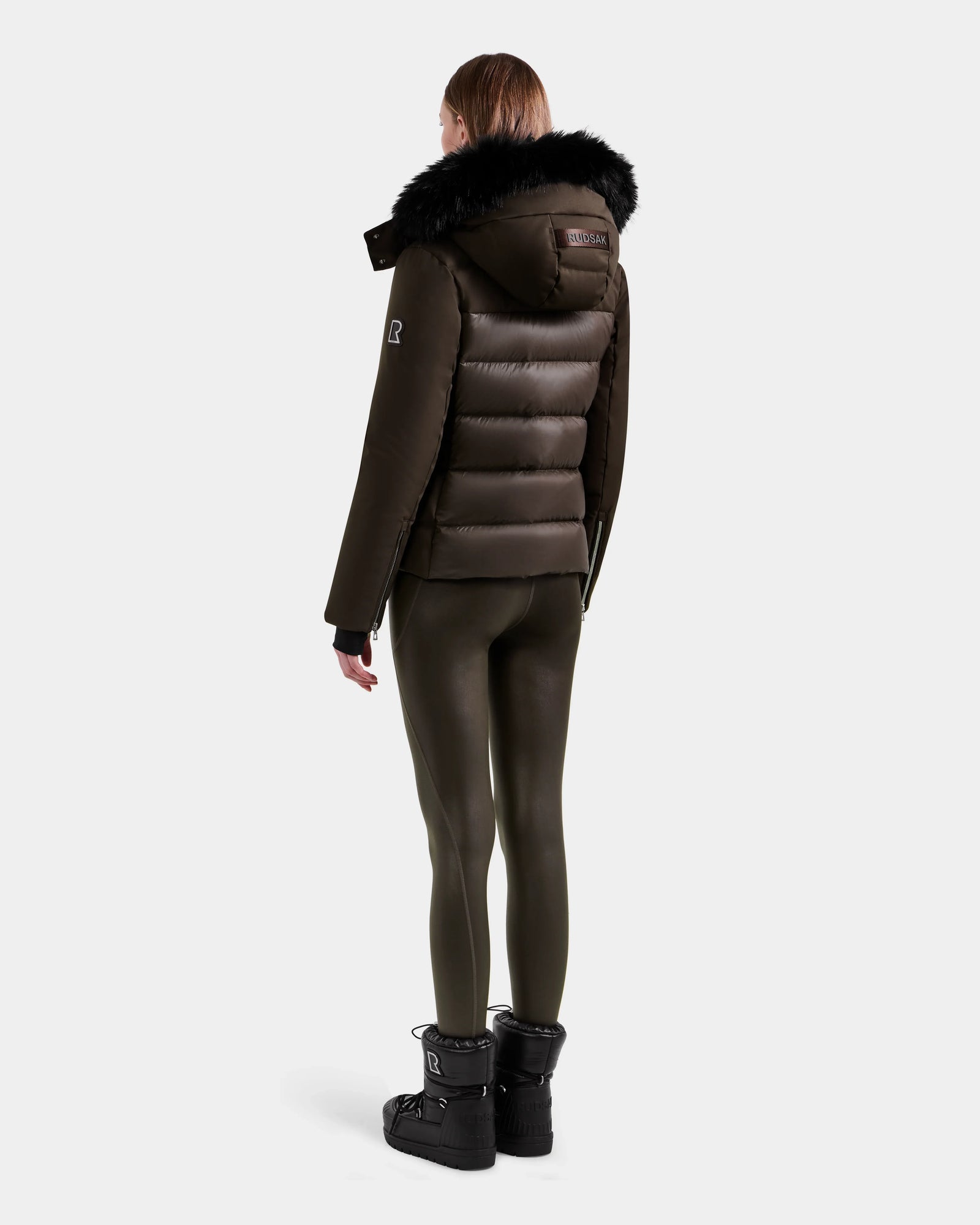 A person stands facing away, wearing the Rudsak MIRRY M - L124533 Espresso women's quilted down coat with a fur-trimmed hood, dark shiny leggings, and black winter boots against a plain white background.