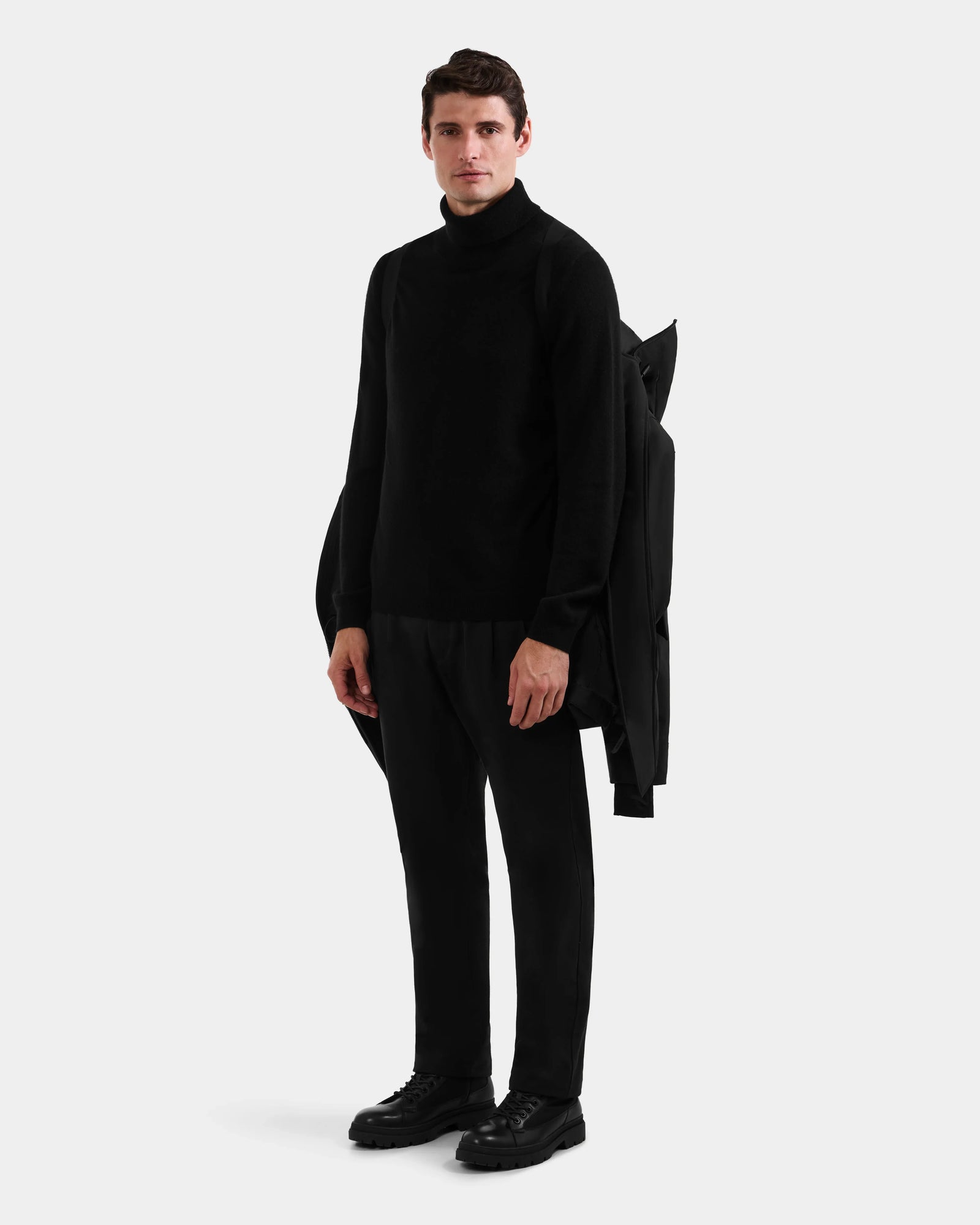 A man faces forward against a plain light gray background, wearing the Rudsak MITCHELL - M125513 Black turtleneck sweater, black pants, and shoes, with a winter parka draped over his left shoulder.