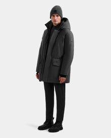 A person stands facing forward in the Rudsak MITCHELL - M125513 Grey men’s down parka, crafted with RDS-certified down, paired with black pants, black boots, and a gray knit beanie against a plain light background.