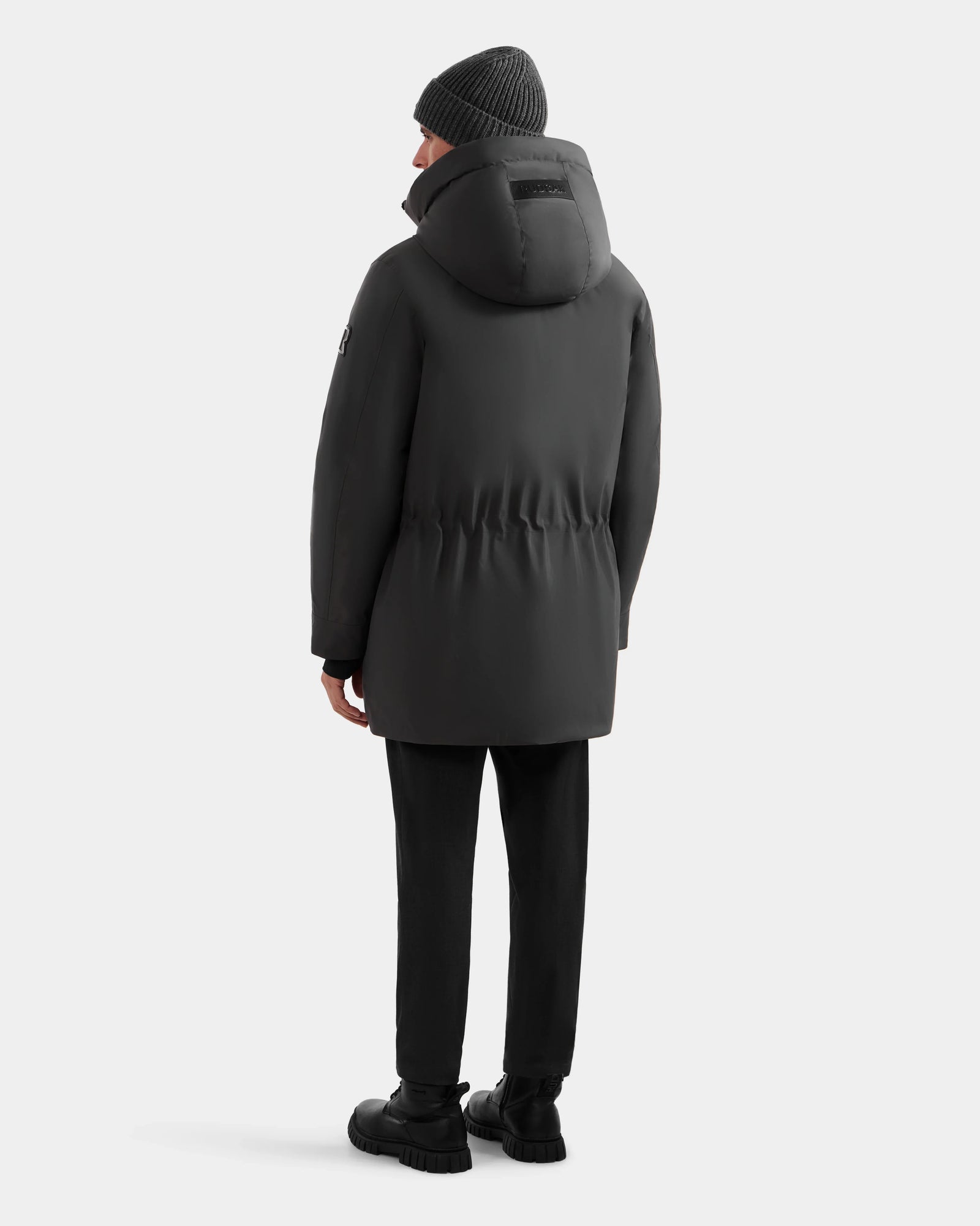 A person in the Rudsak MITCHELL - M125513 Grey men’s down parka stands facing away from the camera, dressed in black pants, black boots, and a gray knit beanie against a plain white background—a true winter essential.