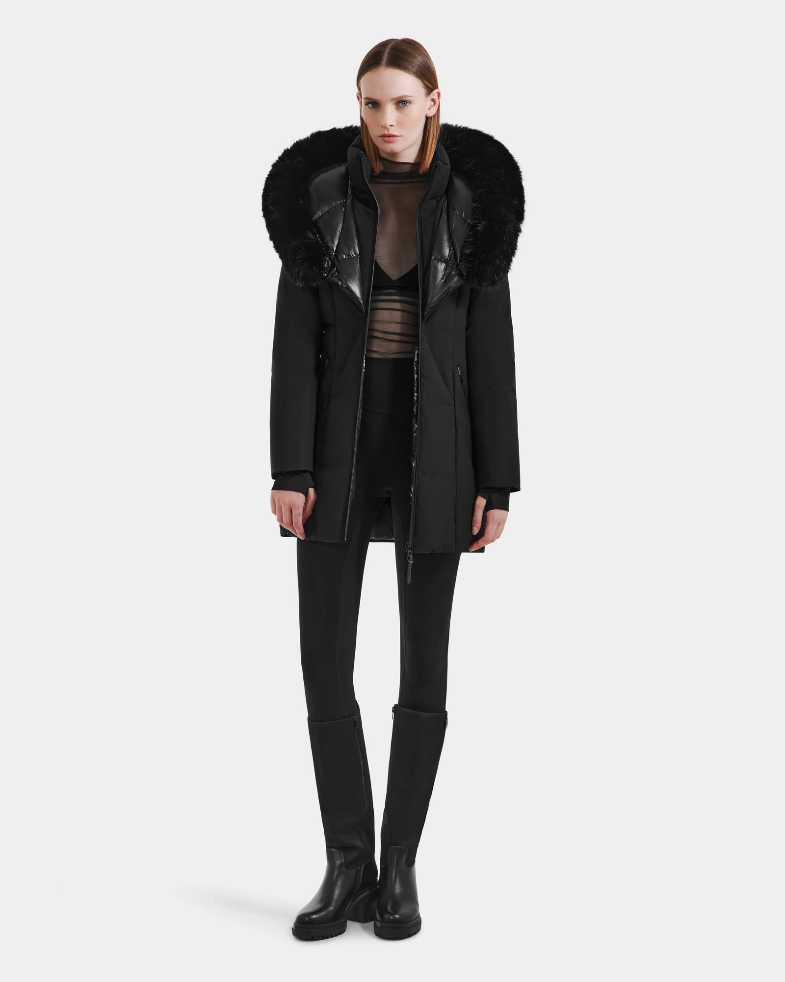 A woman faces forward against a white background, wearing the Rudsak MODA M - L125555 Black winter coat with a large fur-trimmed hood, styled with a sheer black top, black pants, and knee-high black boots.