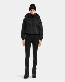 A woman stands facing forward wearing the Rudsak OPRAH - L125546 Black women’s cropped down puffer with RDS-certified down, styled with black leggings, boots, and a knit beanie against a plain light background.