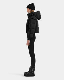 A woman in profile wears the Rudsak OPRAH - L125546 Black women’s cropped down puffer with RDS-certified down, styled with a black beanie, leggings, and winter boots against a plain light backdrop.