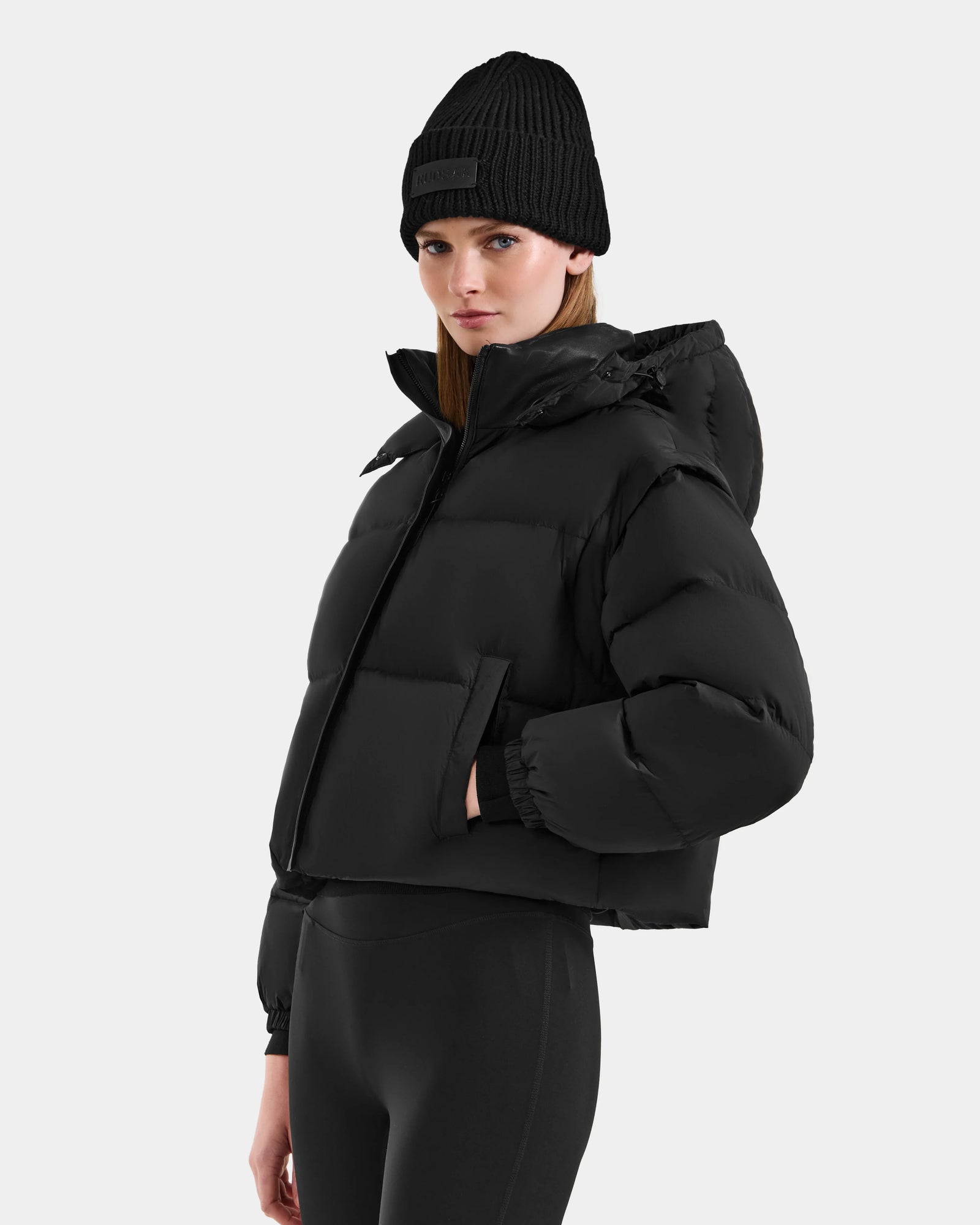 A woman in the Rudsak OPRAH - L125546 Black women’s cropped down puffer, paired with a black beanie and leggings, stands against a plain light background, looking to the side with one hand in her pocket.
