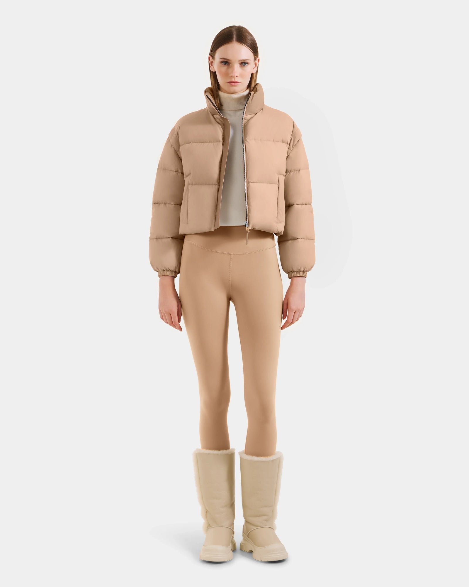 A woman faces forward, wearing the Rudsak OPRAH - L125546 Vanilla cropped down puffer with removable sleeves, matching beige leggings, a light turtleneck, and tall beige fur-lined boots against a plain white background.