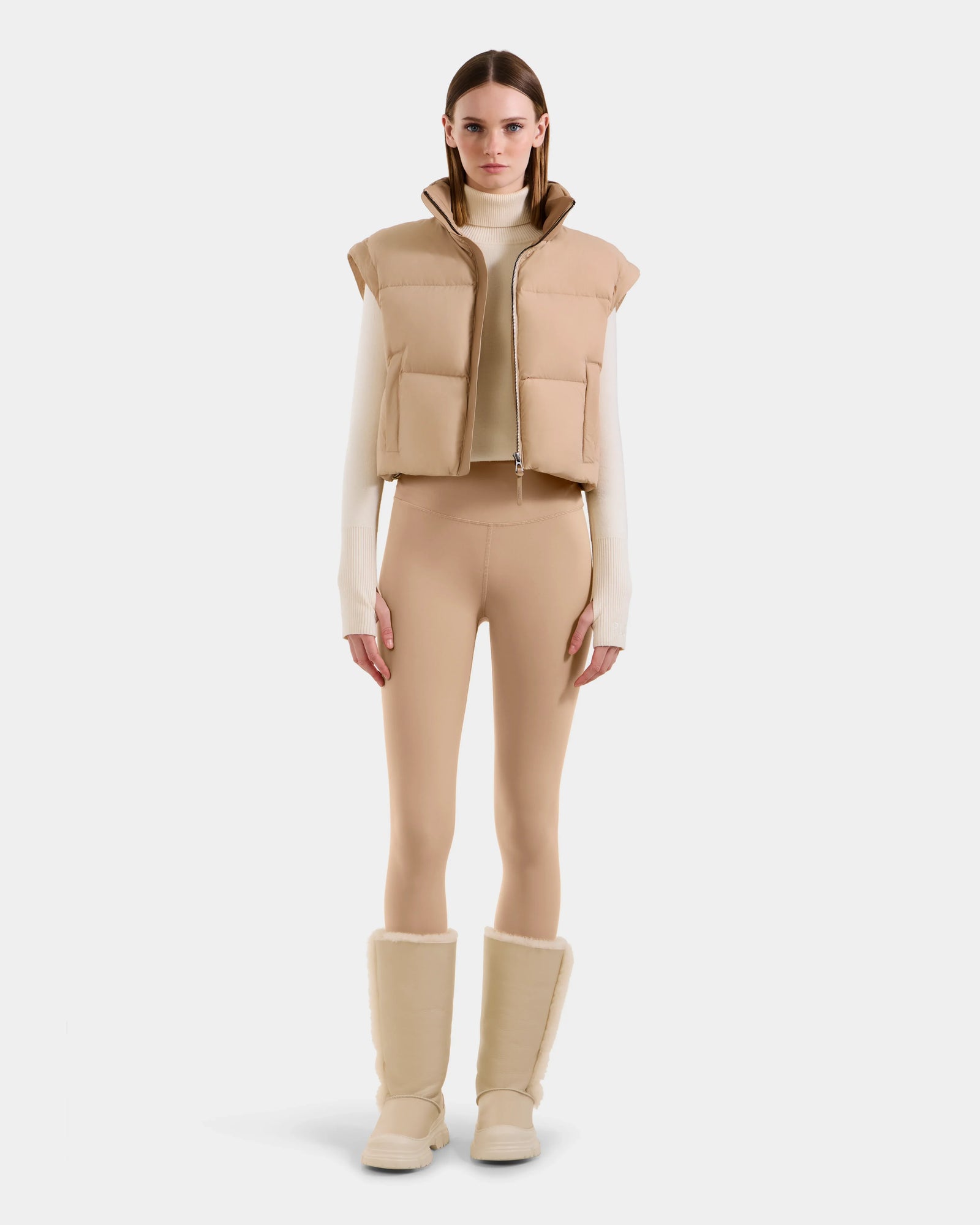 A woman faces forward against a white background, wearing the Rudsak OPRAH - L125546 Vanilla women's cropped down puffer with removable sleeves, paired with a cream turtleneck, tan leggings, and beige shearling boots.