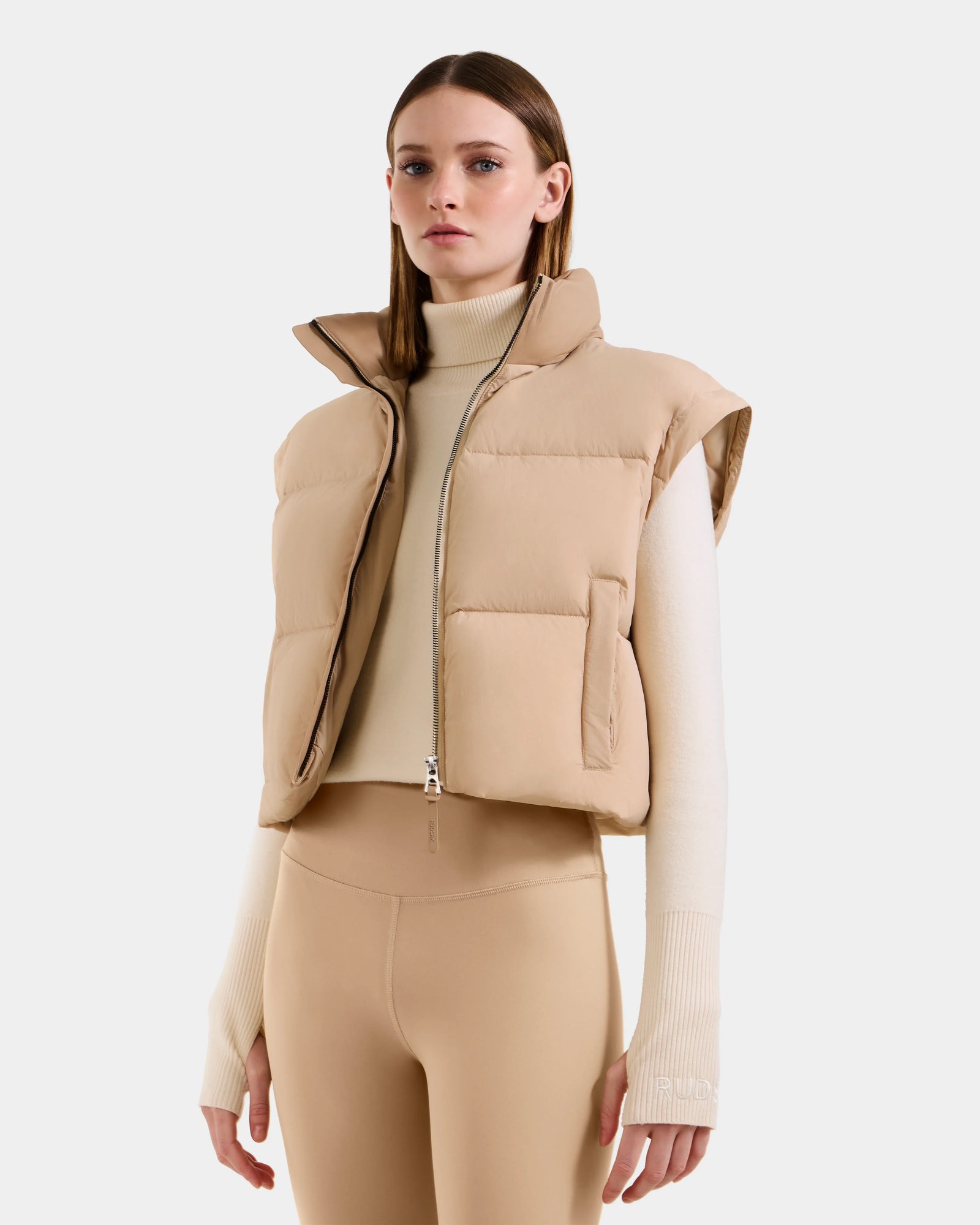 A woman wears the Rudsak OPRAH - L125546 Vanilla women’s cropped down puffer over a cream turtleneck and beige leggings, standing against a white background with a neutral expression.