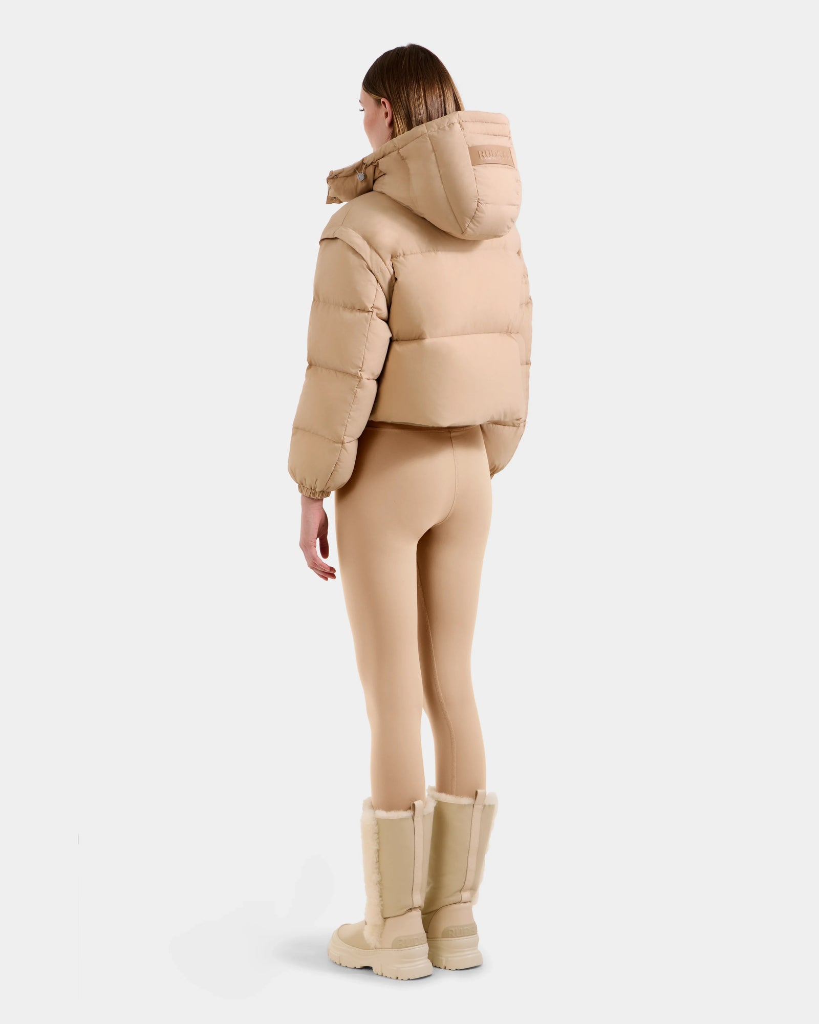 A person stands facing away, wearing Rudsak's OPRAH - L125546 Vanilla RDS-certified women’s cropped puffer jacket with a hood, paired with form-fitting beige leggings and beige knee-high fur-trimmed boots, against a plain white background.