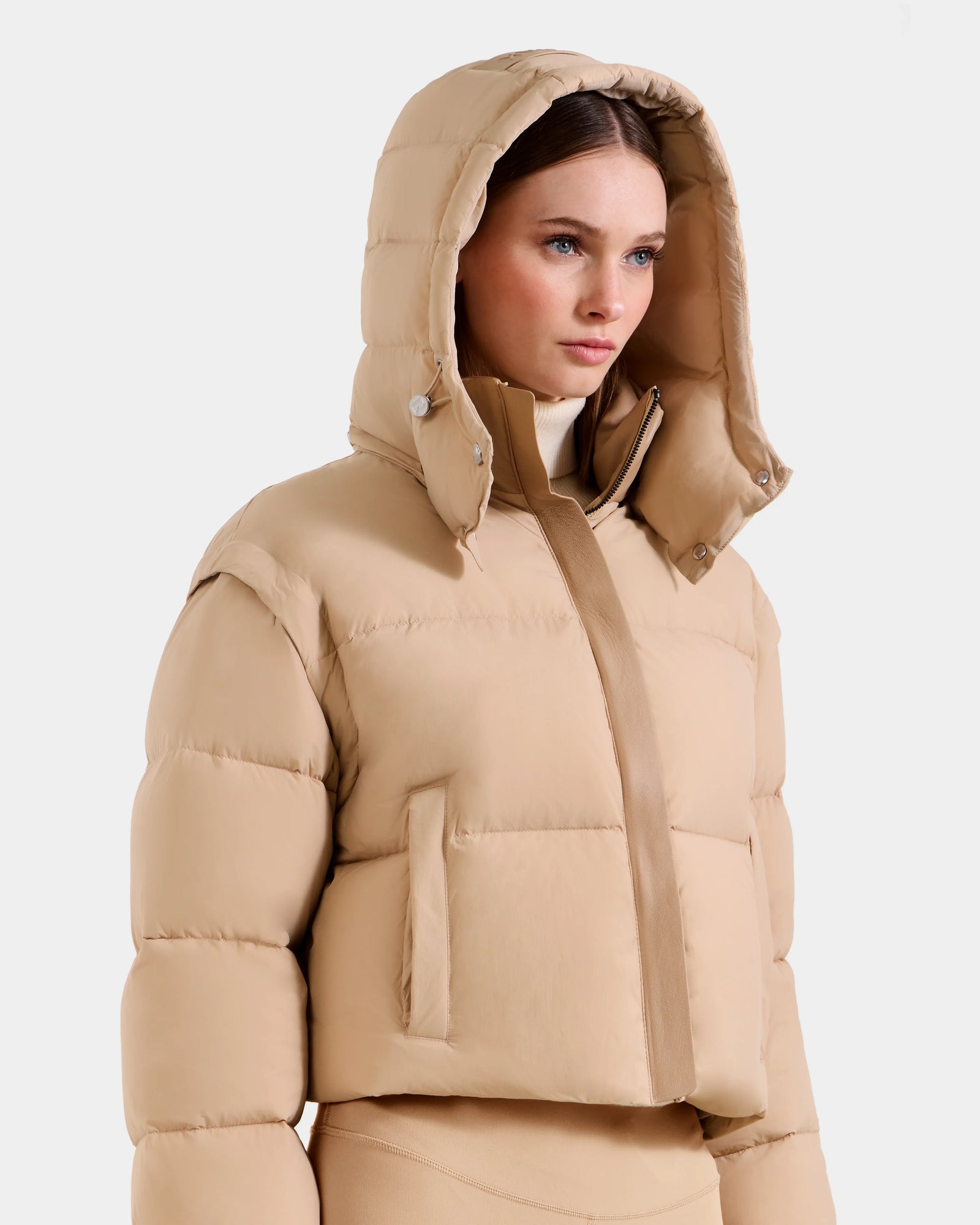 A woman wears the Rudsak OPRAH - L125546 Vanilla puffer jacket with hood, featuring RDS-certified down and removable sleeves for versatile styling, as she stands against a plain white background looking slightly right.