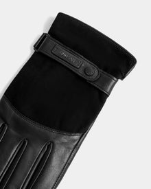 A close-up of the Rudsak PARKER - M425502 Black men’s leather glove, showing its strap with embossed text and a metal snap button, photographed on a light gray background.