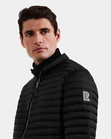 A man with short brown hair wears the Rudsak REYMOND M - M125523 Black down jacket, crafted from recycled nylon with an R logo patch on the sleeve, looking slightly left against a plain light background.