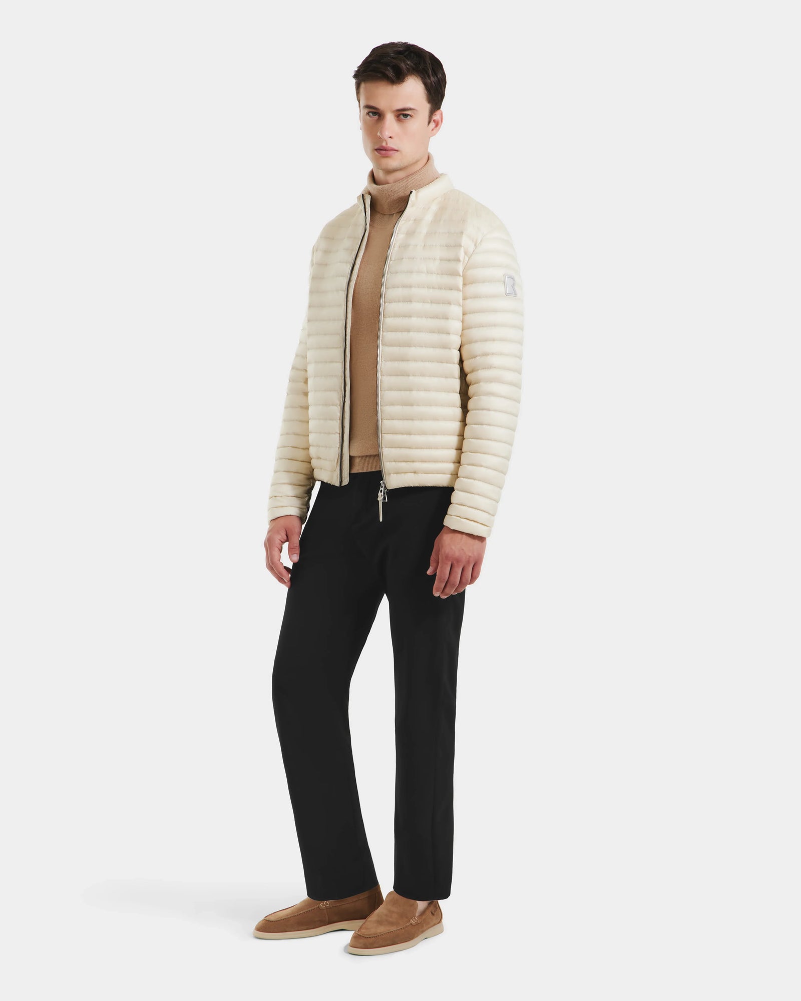 A man wears the Rudsak REYMOND M - M125523 Vanilla down jacket, crafted from recycled nylon, over a beige turtleneck with black pants and tan loafers, standing against a plain white background.
