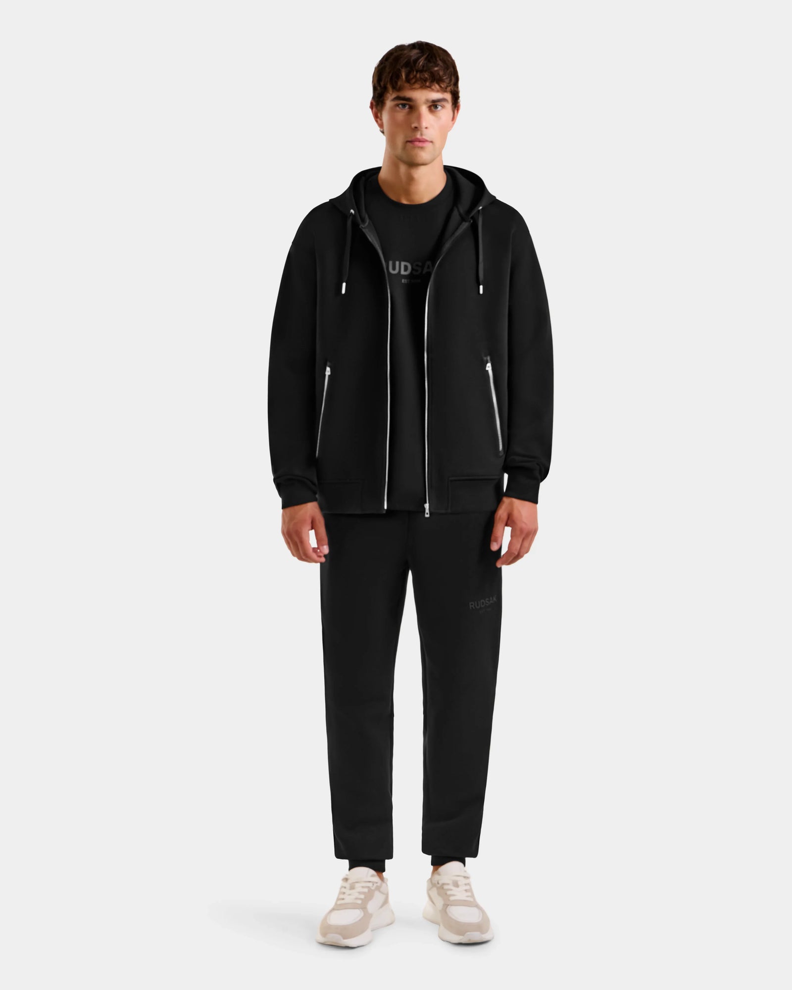 A young man faces forward, wearing the Rudsak ROGER - U523507 Black M unisex zip-up hoodie with black pants, a black T-shirt, and white sneakers, showcasing effortless urban style against a plain light background.