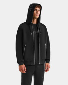 A man wears the Rudsak ROGER - U523507 Black M hoodie with silver zippers over a black shirt and matching black pants, standing against a plain light gray background—a sleek look in urban fashion.