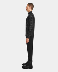 A man stands in profile against a plain background, wearing the Rudsak ROMEO - 6524525 Black stretch active top—a black patterned, long-sleeve, mock-neck zip-up—paired with black pants and lace-up boots.