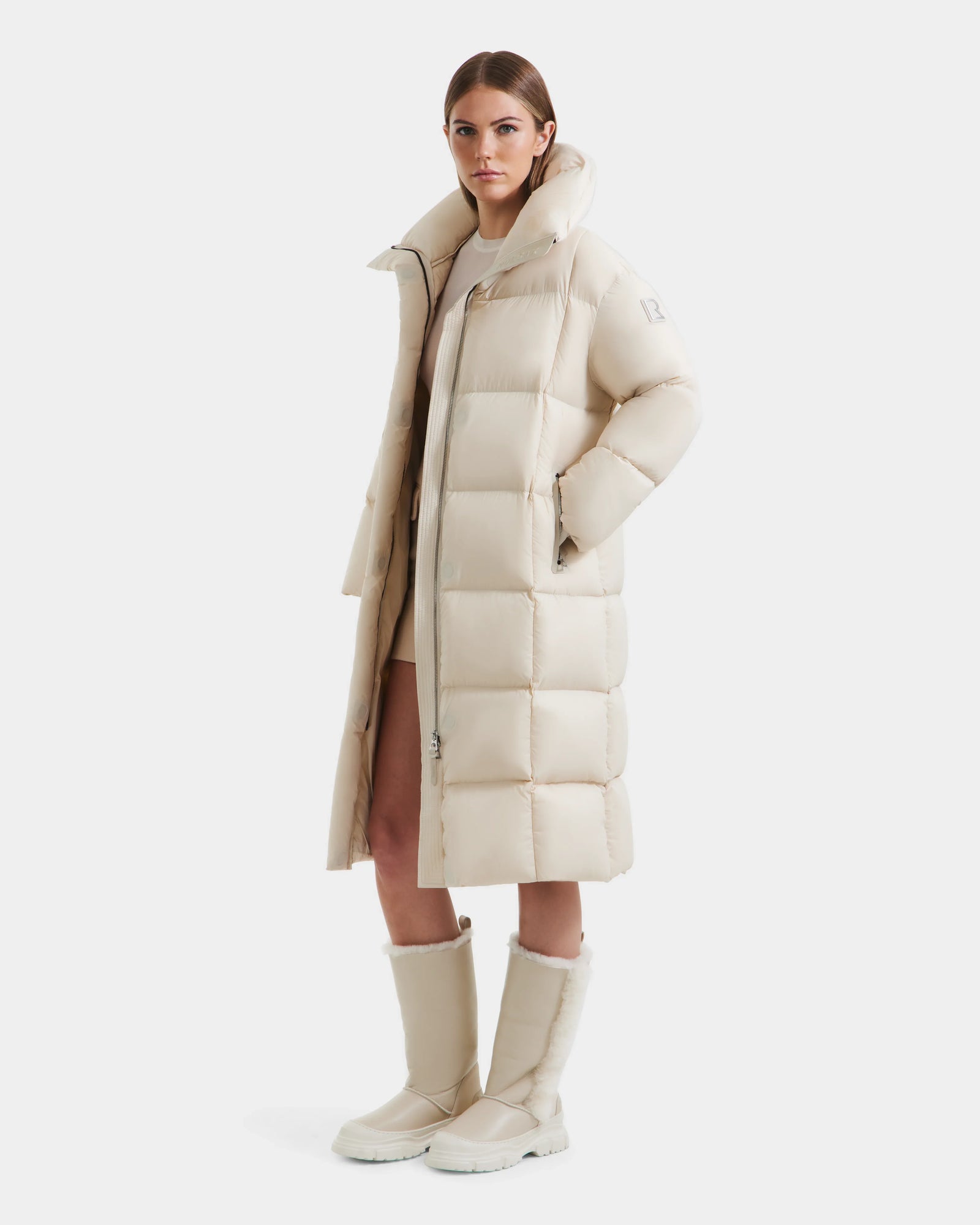 A woman stands facing forward in the Rudsak SASHA - 8125686 Vanilla long down puffer coat with large quilted sections, wearing matching off-white boots and her hands in her coat pockets, against a plain white background.