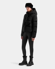 A person wearing the Rudsak STEFANIE M - L125526 Black puffer jacket with a hood, black leggings, winter boots, and a fur hat stands against a plain white background.