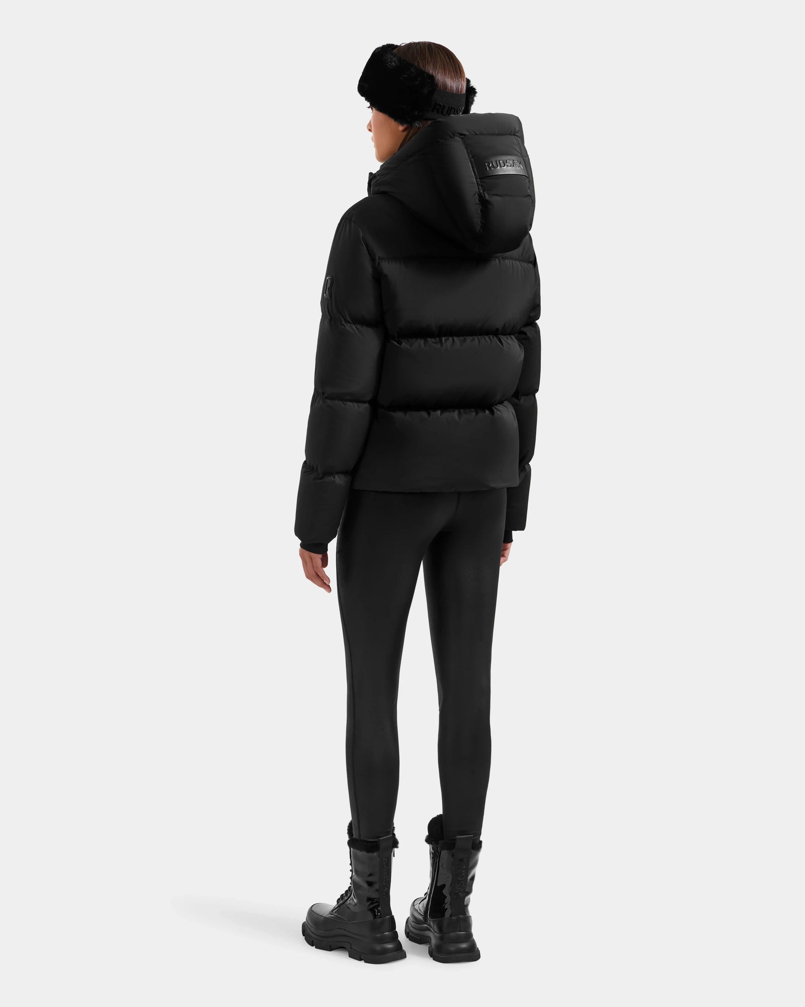 A person faces away, wearing the Rudsak STEFANIE M - L125526 Black puffer jacket with hood, black leggings, combat boots, and a black fur headband against a plain light background.