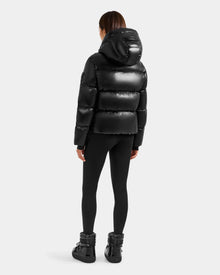 A person with straight brown hair, seen from behind, wears the Rudsak STEFANIE S - L125585 Black shiny puffer jacket with hood, black leggings, and black winter boots against a plain light gray background.