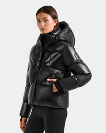 A woman wearing the Rudsak STEFANIE S - L125585 Black shiny quilted down puffer jacket with removable hood and black leggings stands against a plain gray background, glancing to the side with one hand in her pocket.