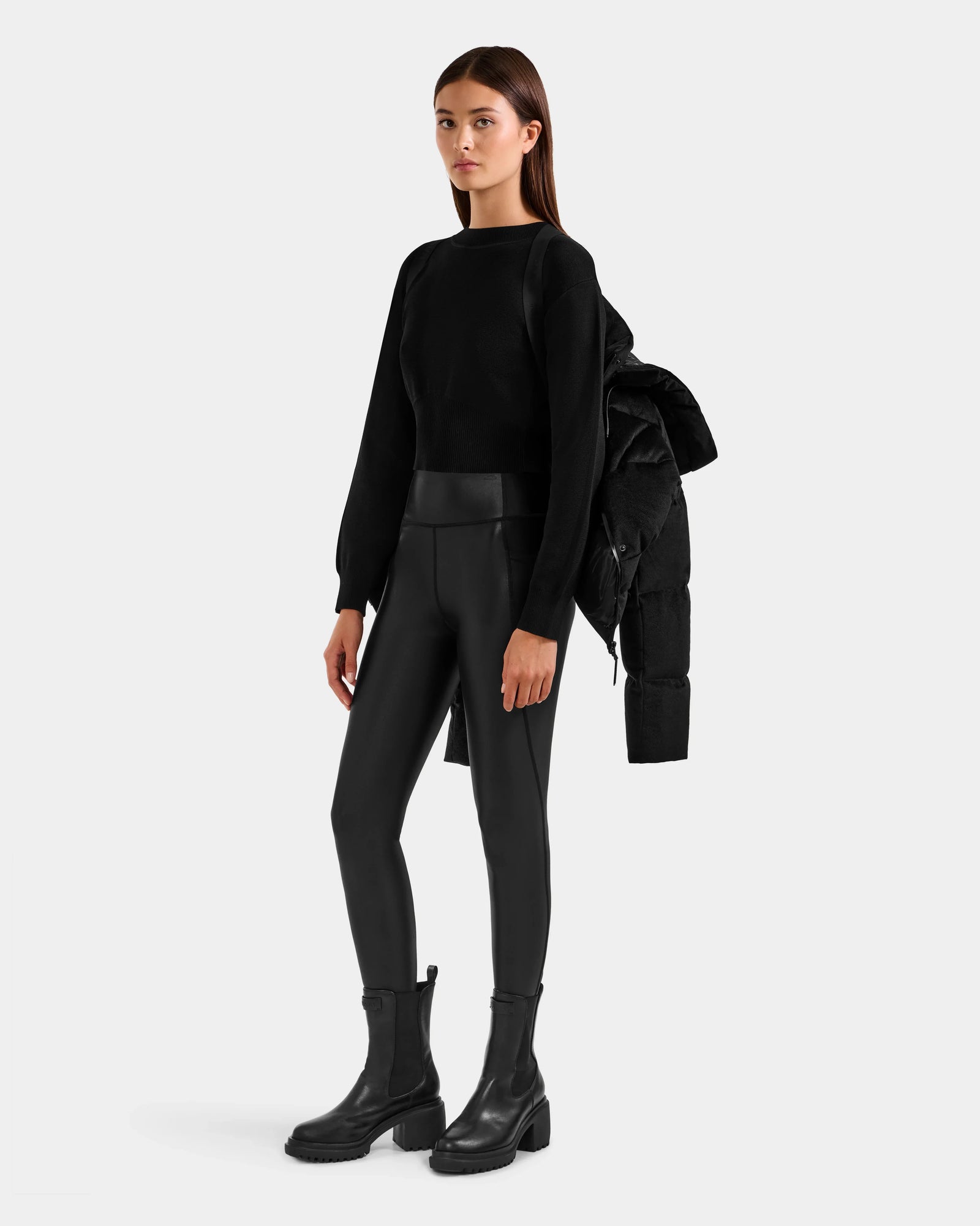 A woman stands facing forward in a black Rudsak STEFANIE V - L125584 down puffer winter jacket draped over one shoulder, styled with black long-sleeve top, faux leather leggings, and chunky boots against a light background.