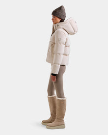 A person stands in profile wearing the Rudsak STEFANIE V - L125584 Vanilla down puffer jacket, paired with taupe leggings, tall beige shearling boots, and a knit beanie against a plain white background.