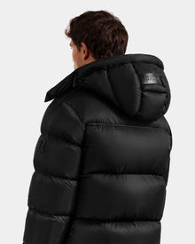 A person wearing the Rudsak STEFANO - 6125574 Black men’s down puffer with hood, seen from behind and slightly to the side, against a plain light gray background.