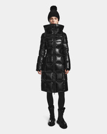 A person wears the Rudsak VENICE S - L124514 Black long, shiny black quilted down puffer coat with a black knit beanie, black leggings, and boots, standing against a plain white background.