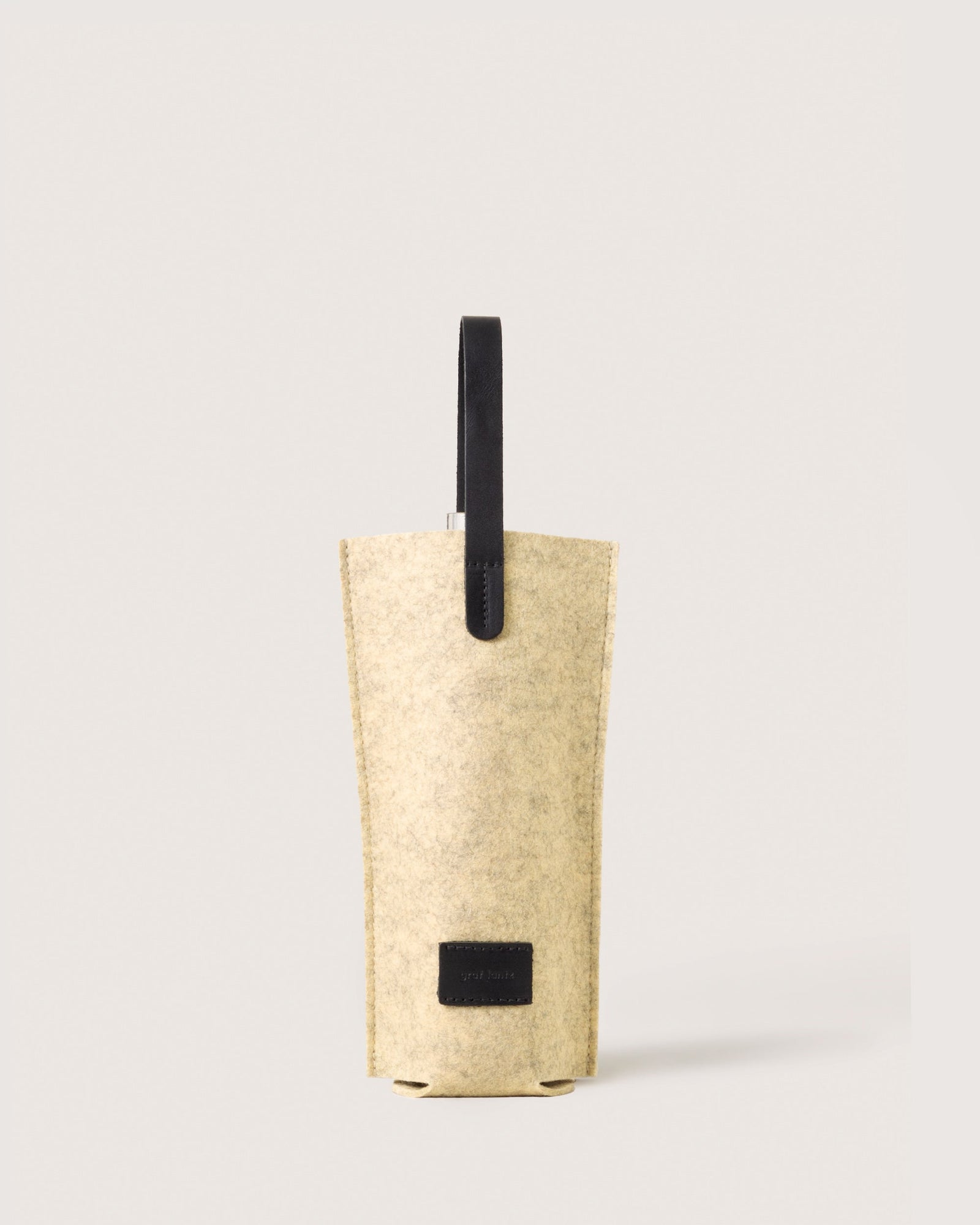 The graf lantz Hana Bottle Bags collection includes a minimalist beige felt wine tote made from Merino wool, featuring leather handles and a small black label near the base, all set against a plain background.