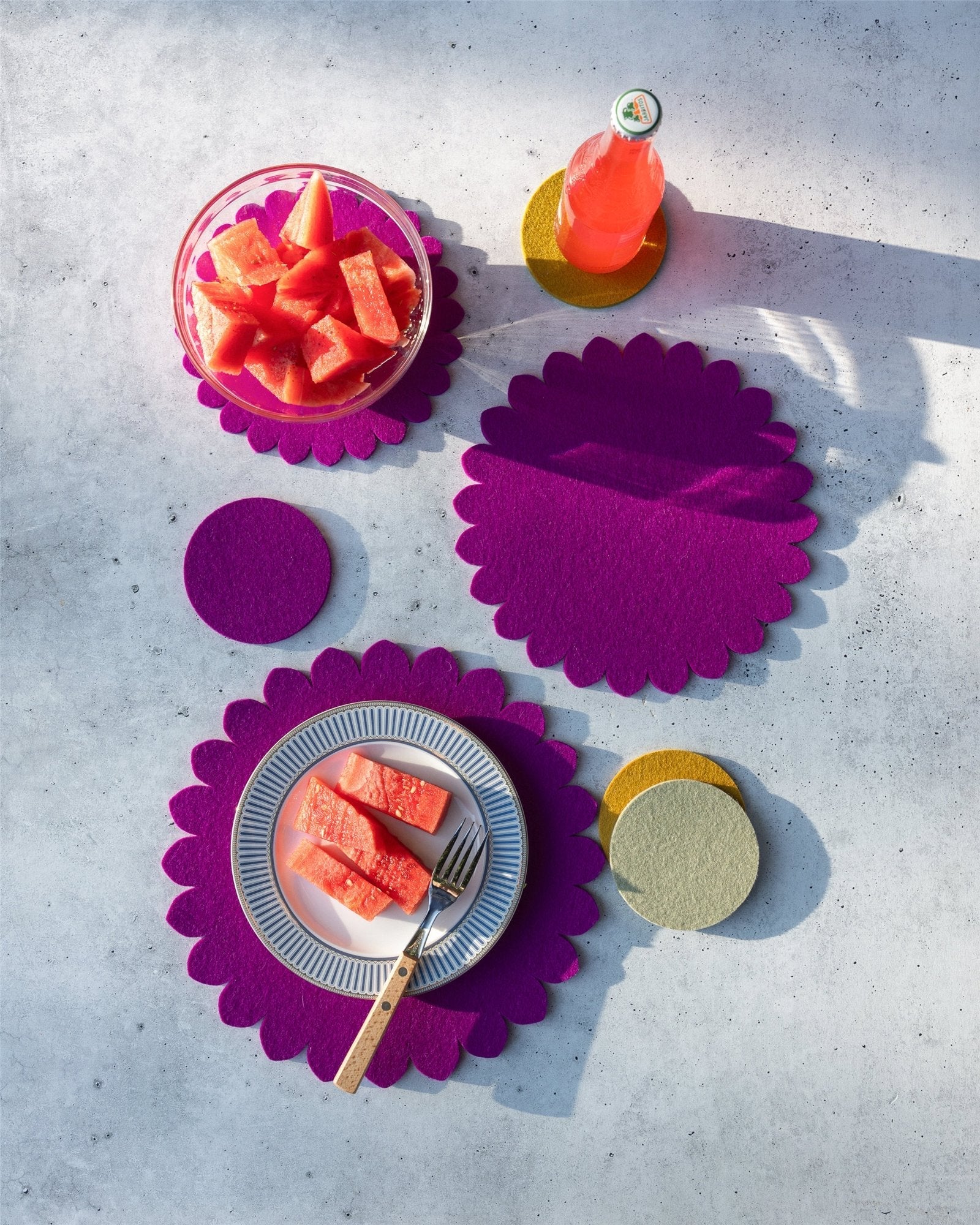 A plate with watermelon and a fork sits on a purple graf lantz Merino wool trivet, beside a bowl of watermelon and a pink drink in a bottle on colorful graf lantz coasters, all arranged on a light surface with soft shadows.