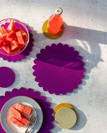 A bowl and a plate with sliced watermelon sit on vibrant pink and yellow scalloped placemats. A bottle of pink drink rests on a graf lantz Merino wool trivet, casting a long shadow for stylish kitchen protection.