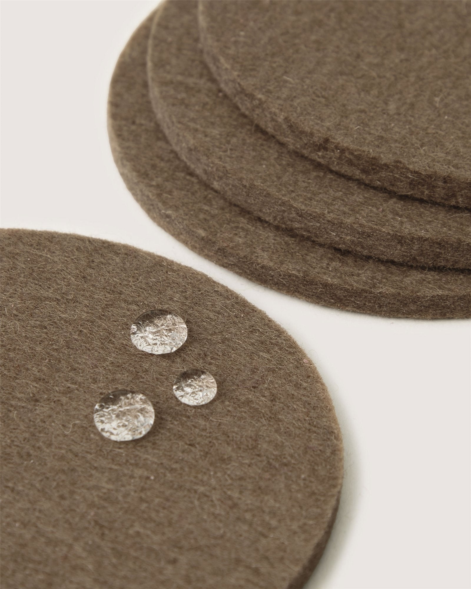 Close-up of round brown Graf Lantz Merino wool coasters on a white surface, with three water droplets beading on the front coaster, highlighting its water-resistant feature—a stylish, sustainable home decor choice.