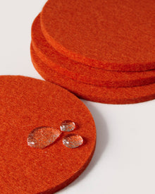 Graf lantz offers stacked round orange felt coasters made from premium German Merino wool, with a rich texture. Three small droplets glisten on the top coaster, while the plain background highlights the vivid color and quality material.