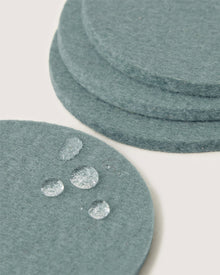 A close-up of teal Merino wool coasters by Graf Lantz, with one in the foreground featuring water droplets—an elegant and sustainable home decor choice.