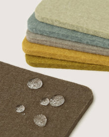 A stack of rectangular Graf Lantz Merino wool coasters in muted colors sits in the background, while a dark brown Graf Lantz German Merino wool coaster with several water droplets is in the foreground.