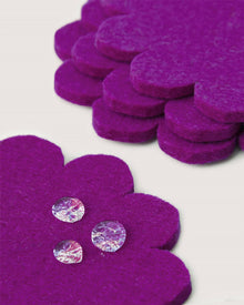 Three clear gemstones sit on a purple, flower-shaped Graf Lantz Merino wool felt coaster, resembling Japanese floral coasters, with a stack of matching coasters providing surface protection in the background on a white surface.