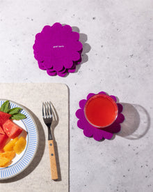A glass of red juice on a gray surface sits on a Graf Lantz Japanese floral coaster, next to a plate of watermelon and orange slices, a fork, and extra stacked purple Graf Lantz coasters for added protection.