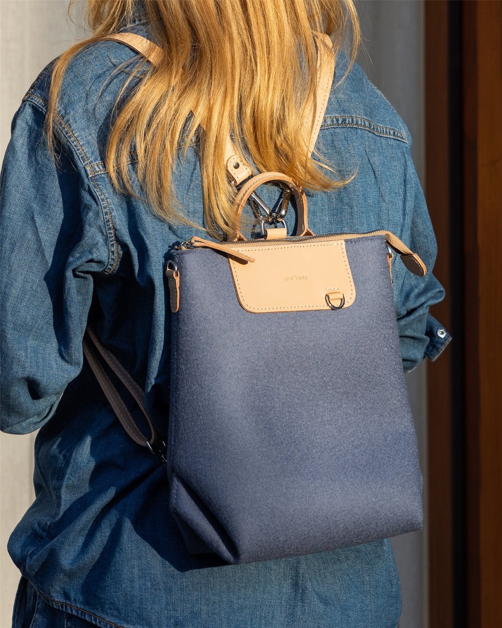 A person with long blonde hair in a denim shirt carries a Graf Lantz backpack—stylish blue with tan accents and leather flap. This eco-friendly fashion piece blends style and sustainability, seen outdoors in sunlit surroundings.