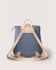 Graf Lantzs blue backpack is showcased from the back, featuring tan leather handles and crossed straps with silver buckles. This multifunctional bag highlights sustainable fashion against a beige background.