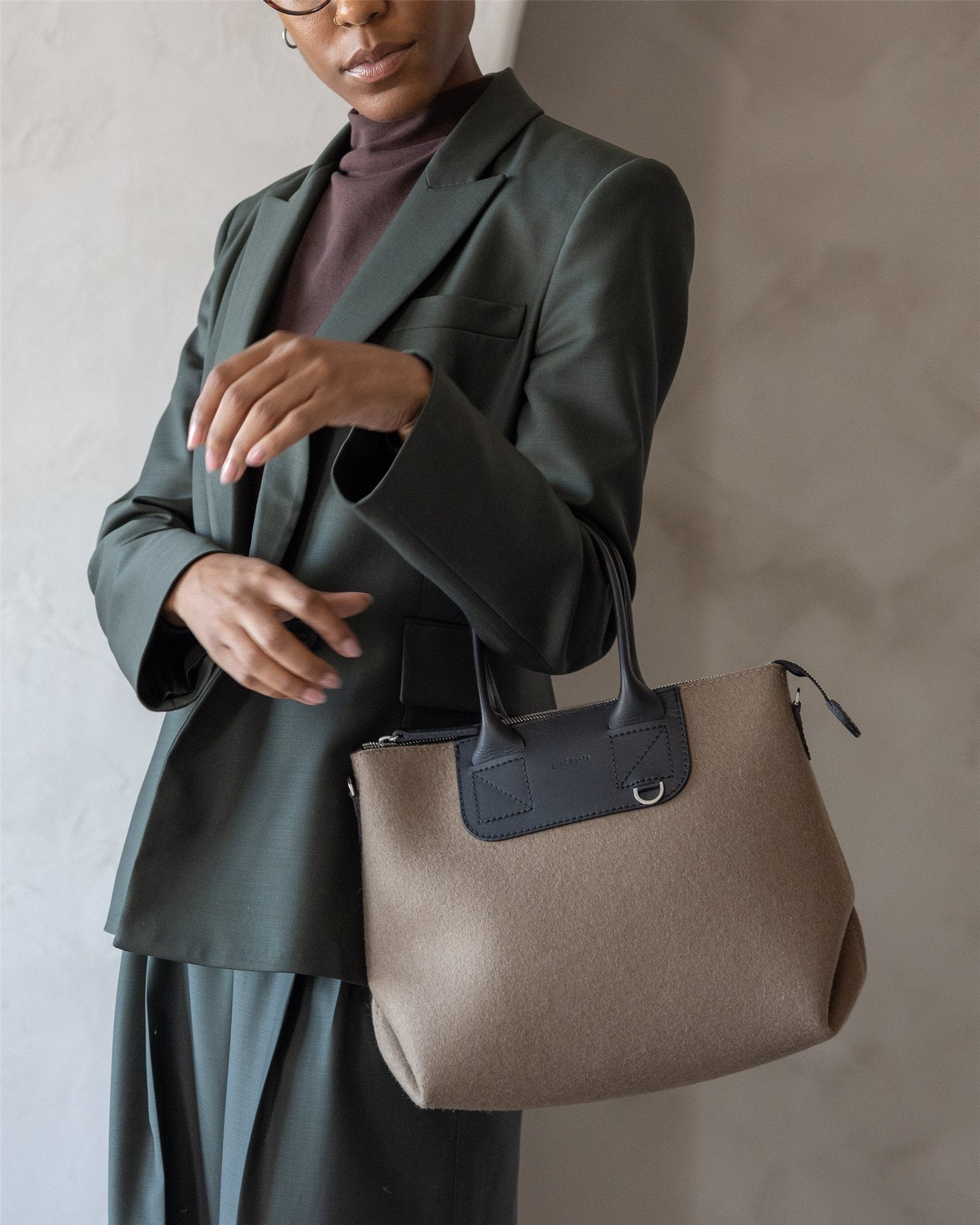 A person in a dark green suit and brown turtleneck poses indoors, holding a beige and black Graf Lantz handbag with short handles against a neutral light gray wall.