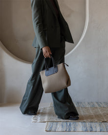 A person in a dark green suit walks indoors on a rug, holding a brown and black Graf Lantz handbag. The curved, textured wall niche is in the background. The person's face is not visible.