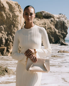 On a rocky beach under a clear sky, someone in a creamy long-sleeved dress holds a Graf Lantz Merino Wool Clutch. Waves gently lap the shore, blending sustainable fashion with the benefits of Merino wool.