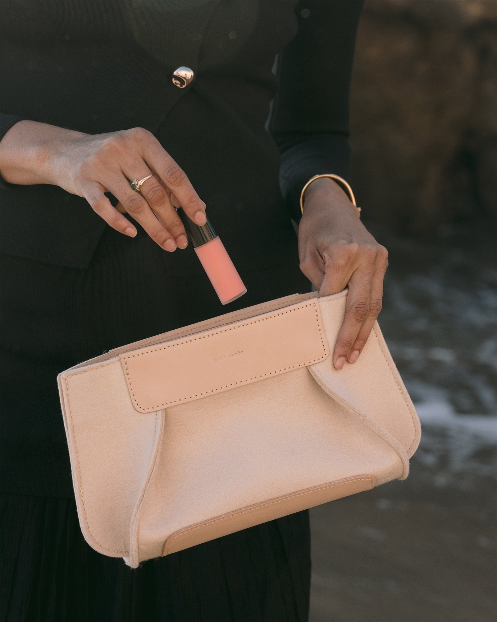 Dressed in black, a person holds a Frankie Merino Wool Clutch by Graf Lantz and a pink lipstick. They sport a gold bracelet and ring, embodying sustainable fashion. The background is blurred, hinting at an outdoor scene.