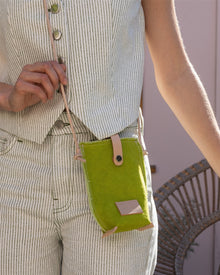 A person in a striped, sleeveless outfit carries the Graf Lantz Hana Phone Crossbody—a green fabric bag with an adjustable strap and button closure—against a wicker chair backdrop.