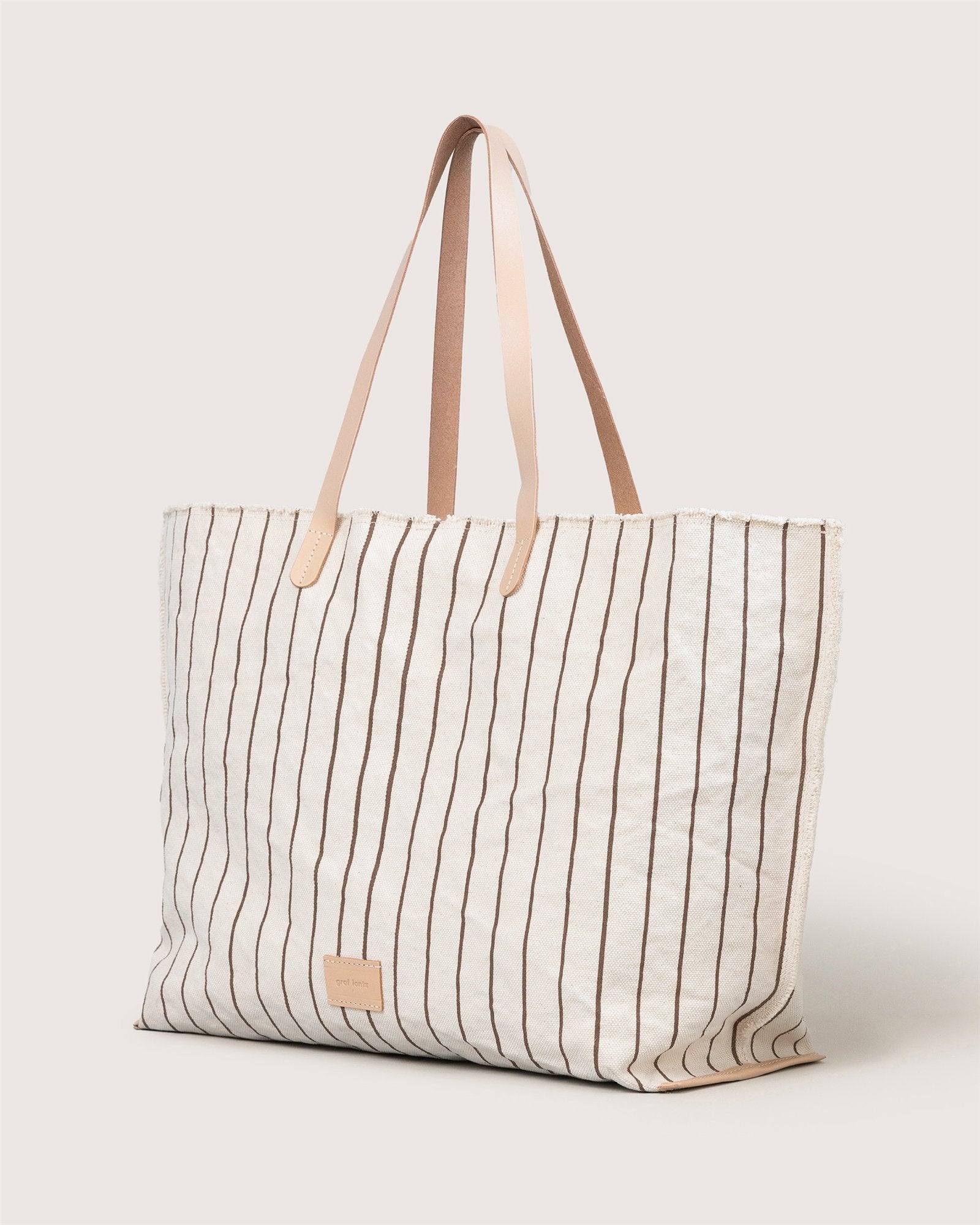 The Graf Lantz Hana Boat Bag is a spacious, cream canvas tote with thin brown stripes and two tan leather handles, shown standing upright against a light background.
