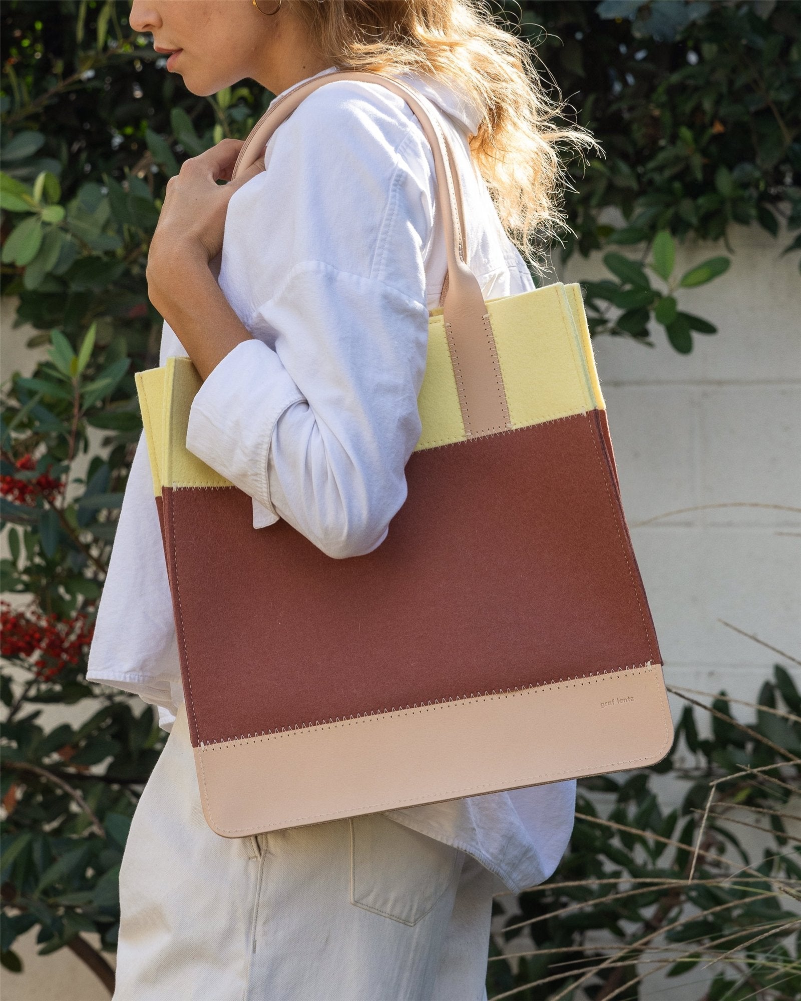 A woman in a white shirt and light pants holds her Graf Lantz Jaunt Midi Tote, featuring beige, brown, and yellow color blocks. Against a leafy green backdrop outdoors, this mid-size carryall blends style with practicality.