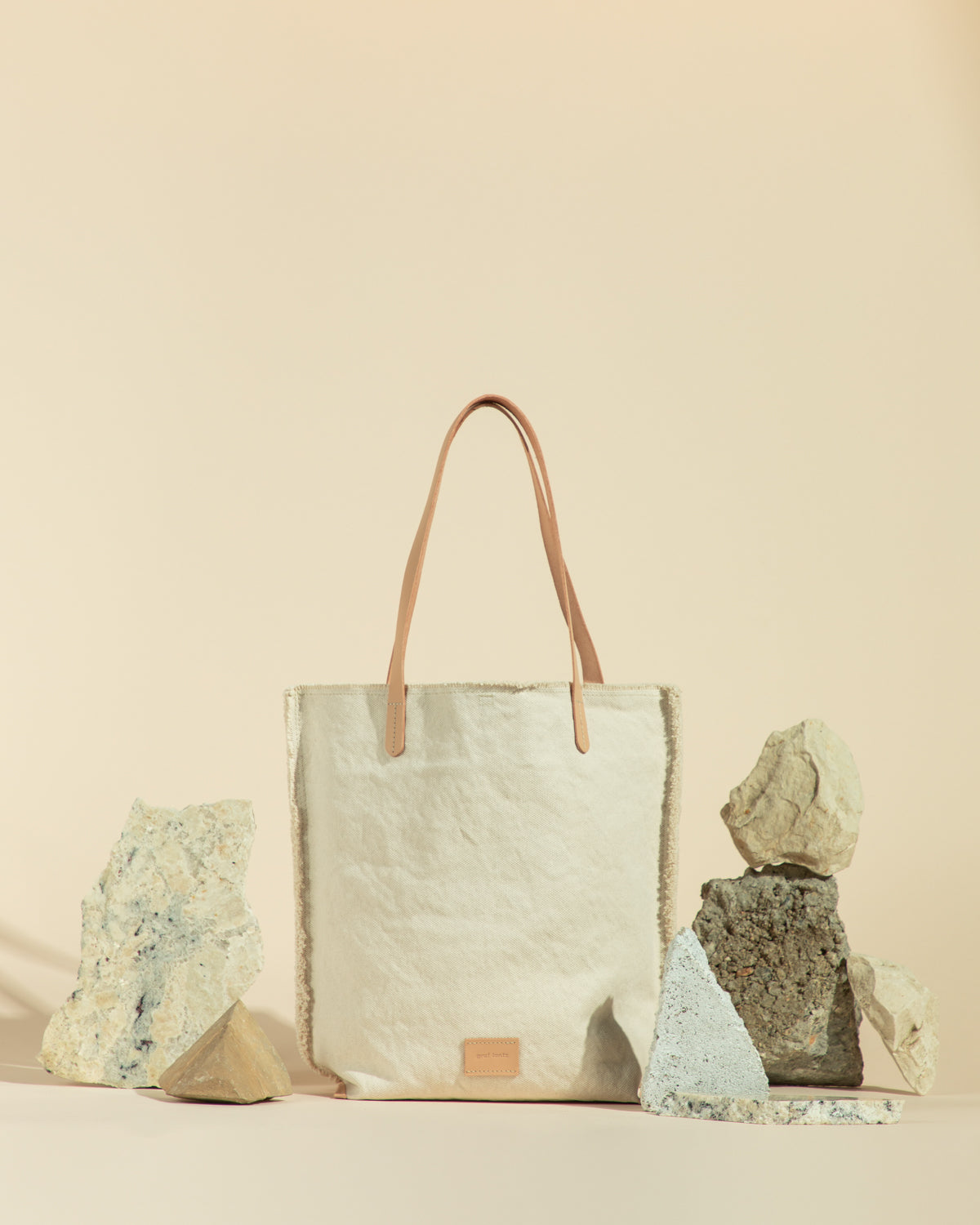 A cream-colored canvas tote bag by graf lantz, made from durable cotton canvas with leather handles, is set against textured stones on a beige background, exuding a minimalist and natural aesthetic.